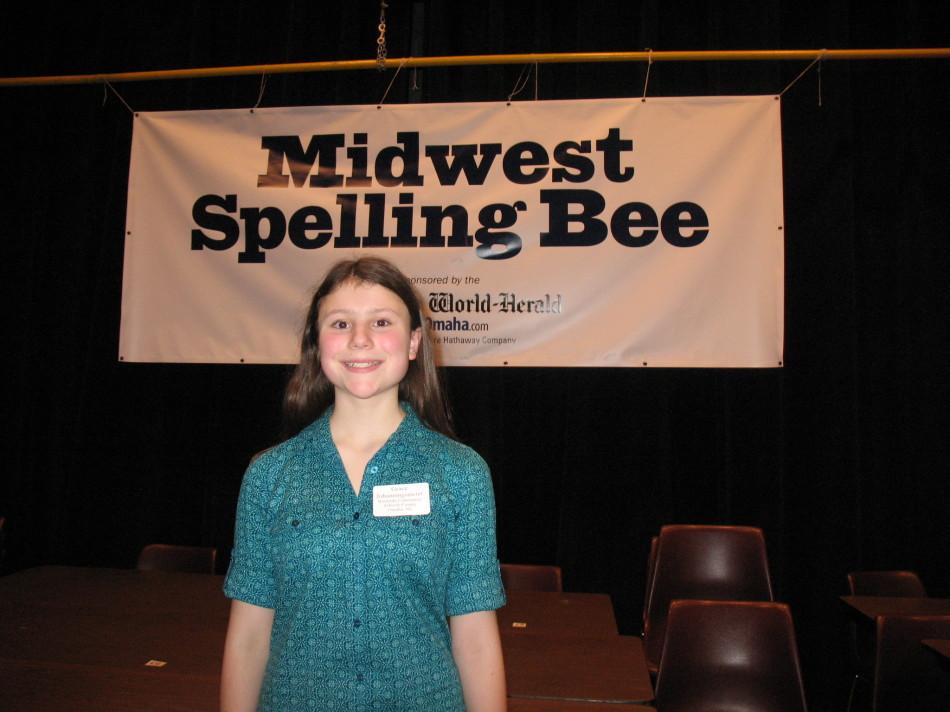 Eighth grader Grace Johanningsmeier stands in front of the banner at the Midwest Spelling Bee. Johanningsmeier won and will compete in the National Spelling Bee in late May.